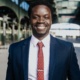 Headshot of Nii Ato Bentsi-Enchill, smiling warmly at camera wearing blue suit, light pink shirt, with red tie w/ small white polkadots, and silver tie bar.
