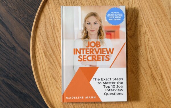 The image shows a hard cover book on a brown round wooden side table. The book is Job Interview Secrets by Madeline Mann