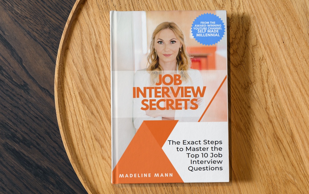 The image shows a hard cover book on a brown round wooden side table. The book is Job Interview Secrets by Madeline Mann