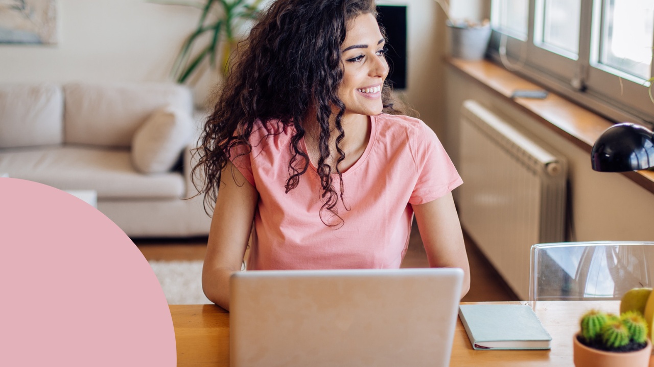 A middle-aged woman with dark hair and a pink t-shirt writes her resume on her laptop. She is preparing to return to work after being a stay-at-home mom.