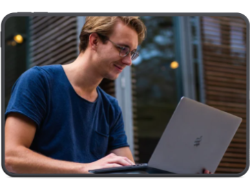 A young man in dark-rimmed glasses sits working on a grey laptop. He is wearing a navy t-shirt and smiles as he updates his resume.